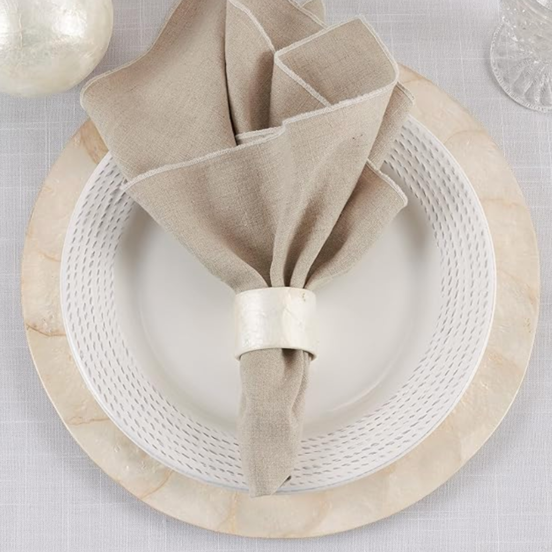 A beige linen napkin with white trim is held by the Saro Trading Capiz Napkin Ring Ivory and rests on a white patterned plate atop a cream charger, with a glass and decorative sphere placed nearby.