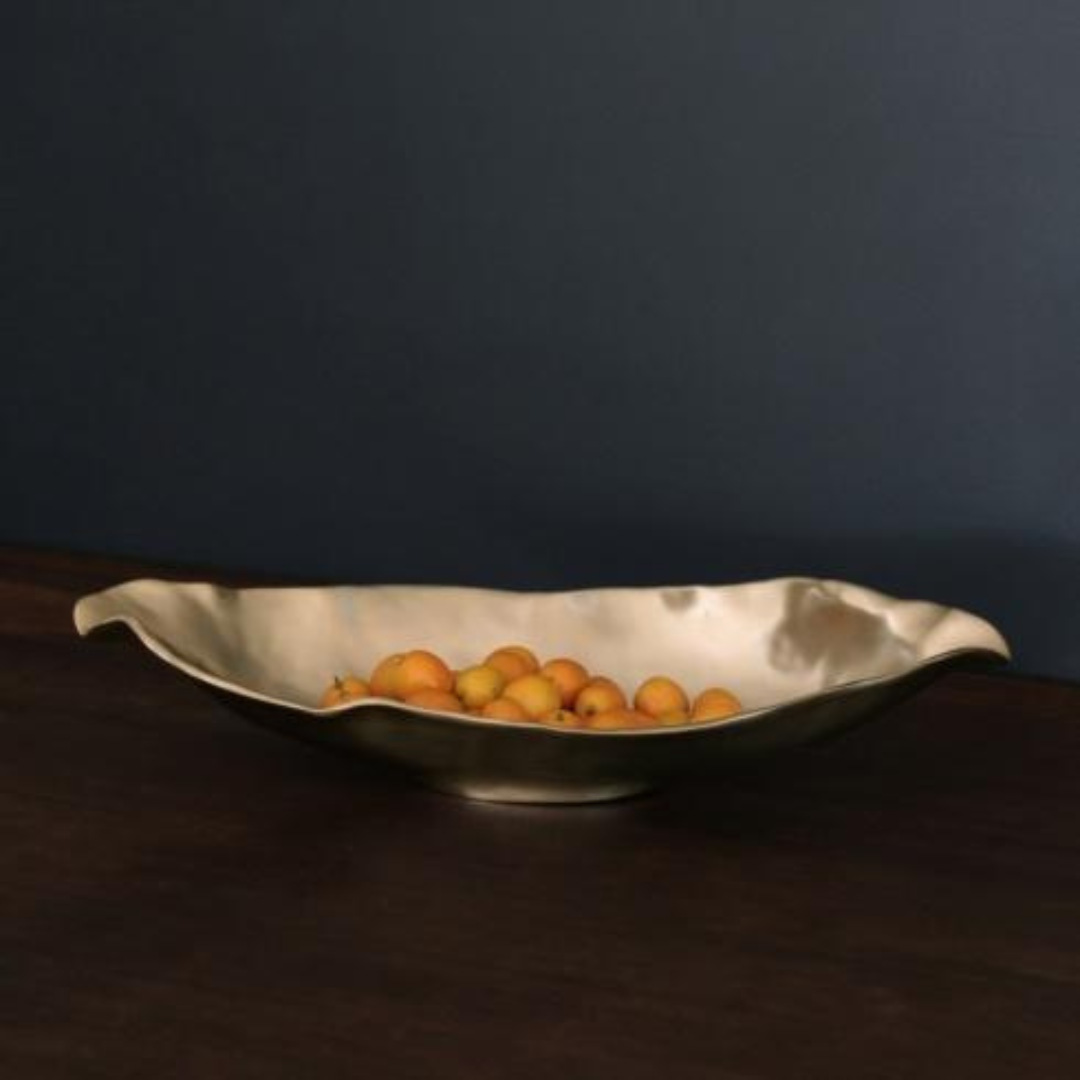 The Beatriz Ball Sierra Maia Long Oval Medium Bowl, a wavy metallic piece, holds small yellow-orange fruits on a dark wood surface set against a matte dark blue wall.