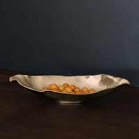 The Beatriz Ball Sierra Maia Long Oval Medium Bowl, a wavy metallic piece, holds small yellow-orange fruits on a dark wood surface set against a matte dark blue wall.