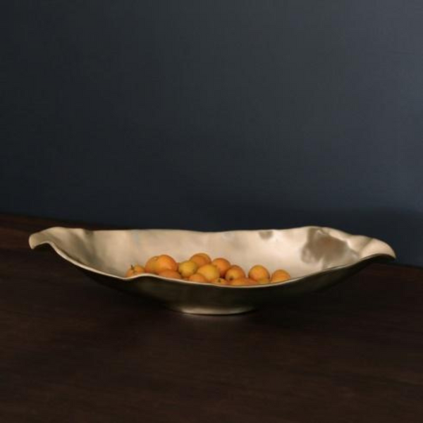 The Beatriz Ball Sierra Maia Long Oval Medium Bowl, a wavy metallic piece, holds small yellow-orange fruits on a dark wood surface set against a matte dark blue wall.