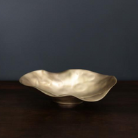 The Sierra Maia Oval Gold Bowl by Beatriz Ball, crafted by hand in a champagne gold finish with wavy edges, adds eco-friendly elegance atop a dark wooden surface against a plain dark gray background.