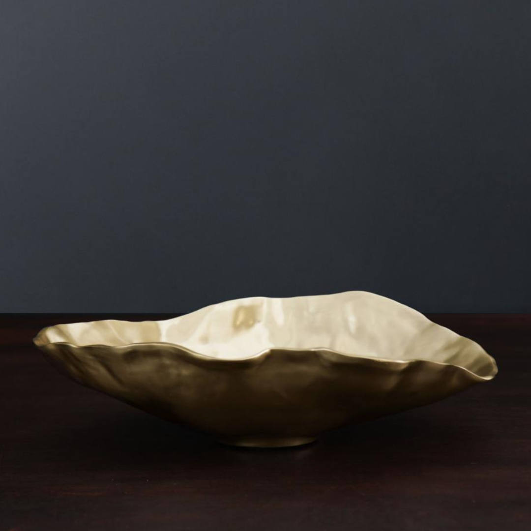 The Beatriz Ball Sierra Maia Oval Gold Bowl, a large handmade aluminum piece in champagne gold, sits on a dark wooden surface against a dark gray background.