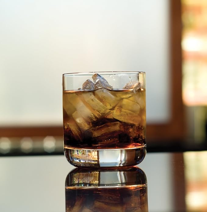 A Zwiesel Fortessa Whiskey Glass from the Simple Set of 6, filled with ice cubes, stands on a reflective surface with its sturdy design illuminated by a softly blurred, warmly lit background.