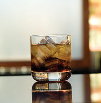 A Zwiesel Fortessa Whiskey Glass from the Simple Set of 6, filled with ice cubes, stands on a reflective surface with its sturdy design illuminated by a softly blurred, warmly lit background.