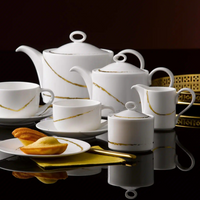 The Sketch Chalk Dinnerware by Royal Crown Derby—a white set with gold accents including a teapot, cups, saucers, sugar bowl, and creamer—rests on a glossy black surface. A plate of madeleines and a yellow napkin complete the scene.