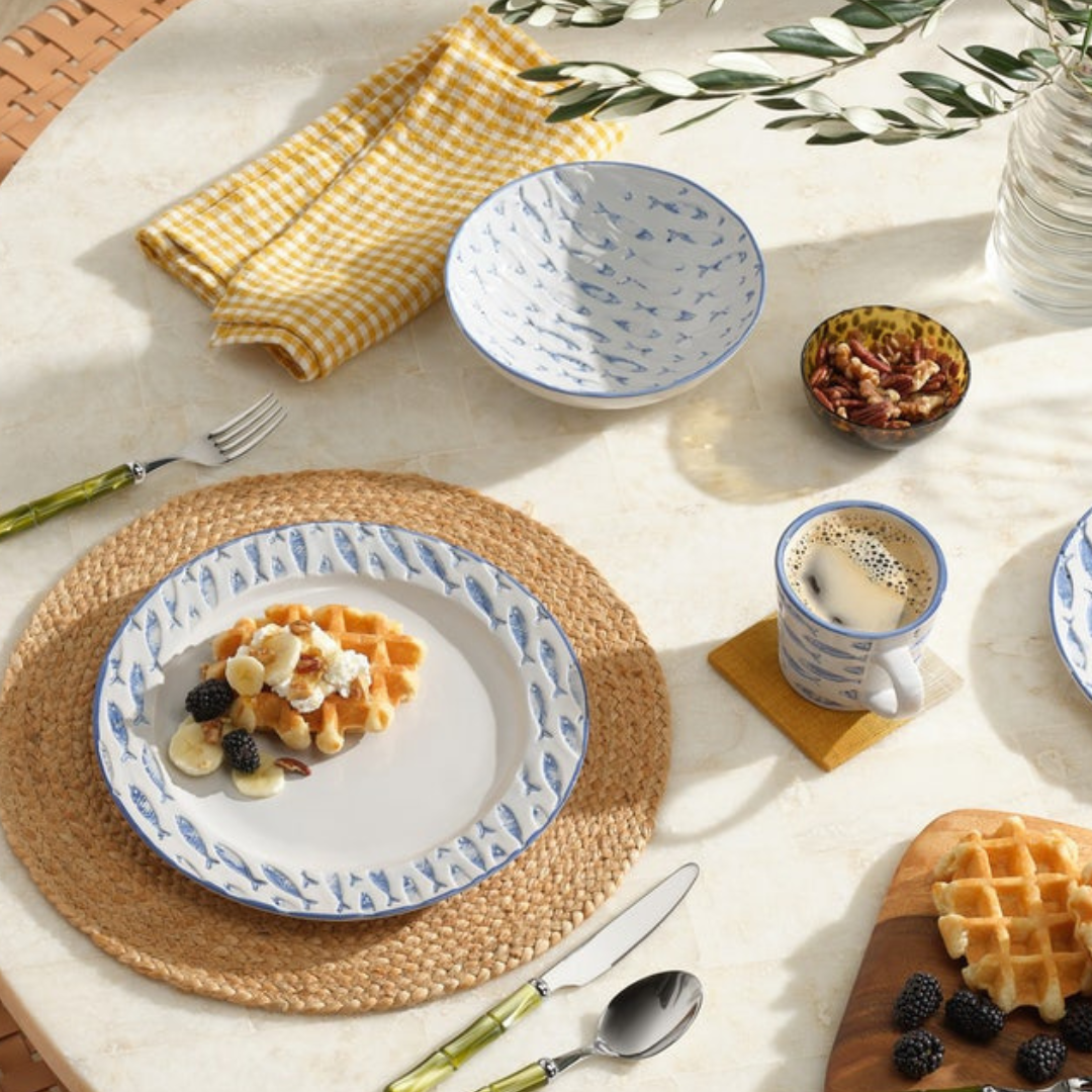 Breakfast table setting featuring waffles with banana slices, blackberries, and pecans, a cup of coffee, the Blue Pheasant Andrew Tortoise Nut Bowl with pecans, yellow gingham napkin, cutlery, and a woven placemat.