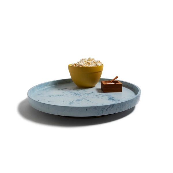 The Trashy Goods Smart Lazy Susan, a round light blue sustainable tray, holds a yellow bowl of popcorn and a small wooden container with spoon, all set on a white background.