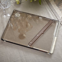 A translucent rectangular Solid Acrylic Challah Board & Knife by Apeloig rests on a beige tablecloth near a wine glass, flowers, and silverware. The tray is labeled “ARLECCHINO COLLECT.”.