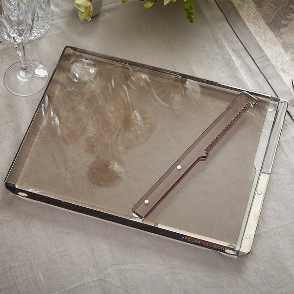 A translucent rectangular Solid Acrylic Challah Board & Knife by Apeloig rests on a beige tablecloth near a wine glass, flowers, and silverware. The tray is labeled “ARLECCHINO COLLECT.”.