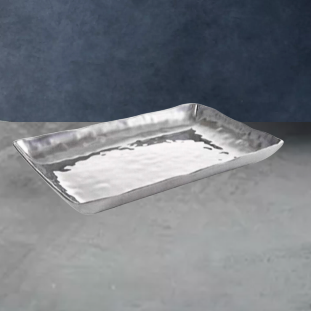 The Beatriz Ball Soho Rectangle Pennsylvania Platter is a silver aluminum alloy tray with a lightly hammered texture and raised edges, perfect as a stylish oven-to-table piece. It is shown on a gray surface against a dark blue background.
