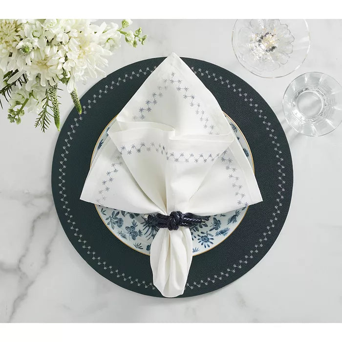 The Kim Seybert Starry Night Napkin Set of 4 in White & Blue, featuring blue star metallic embroidery, is folded and held by a dark blue rope ring on a white plate with blue floral designs atop a dark green placemat.