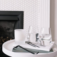 A round white side table holds a Nude Glassware Stem Zero Vertigo Water Glass Set of 2 (lead-free crystal), a frosted candle, books, and black eyeglasses; behind it are a black fireplace and a white hexagon tile wall.