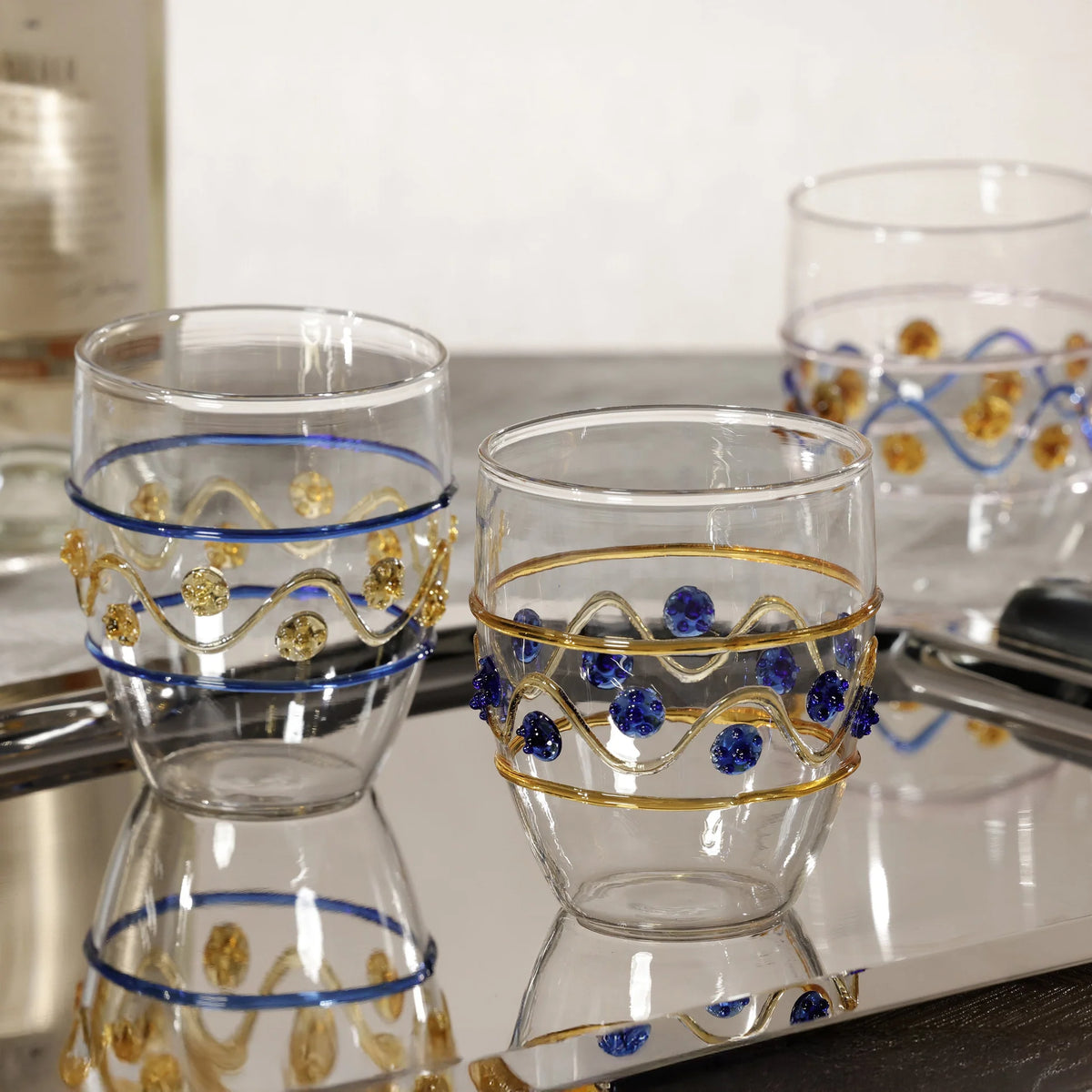 Three Zodax Festa Tumblers in green, amber, and blue tones with wavy metallic designs sit on a reflective silver tray, with a blurred bottle and another stemless glass in the background.