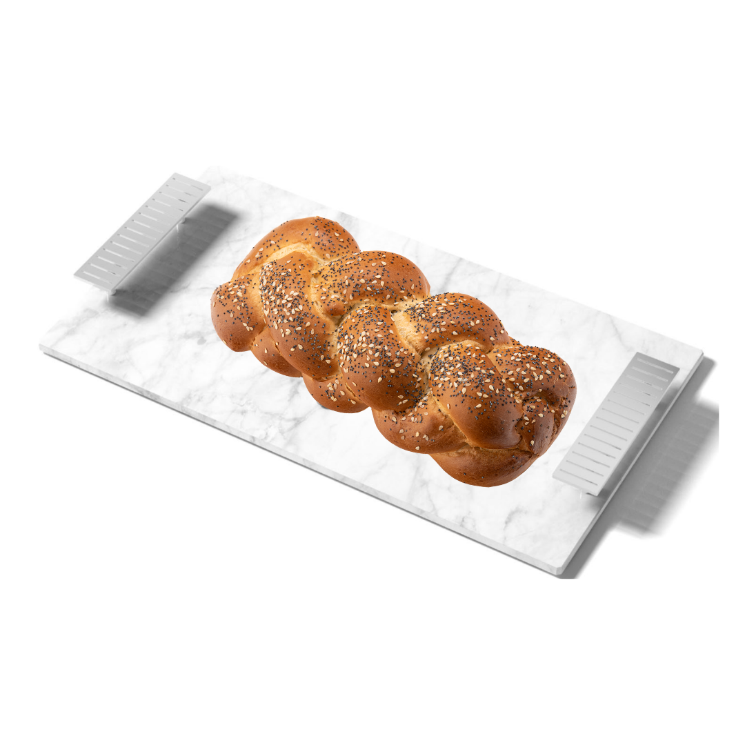 A braided loaf with seeds sits on the Anna New York Striations Cheeseboard & Knife Set, a rectangular marble board featuring silver metal handles at each end.
