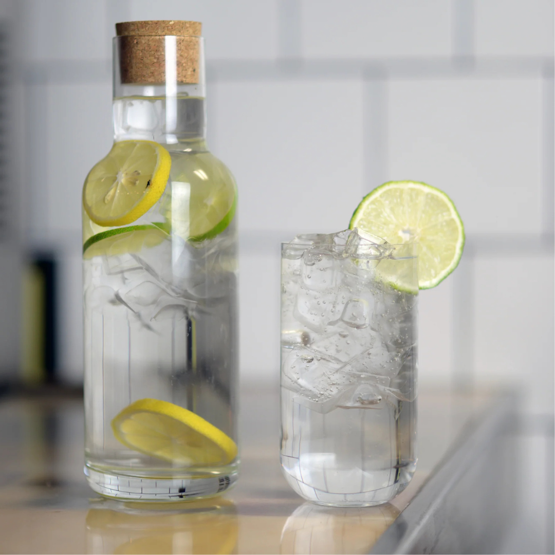 A Luigi Bormioli Sublime Beverage Glass from the Set of 4 and a carafe with cork lid, both filled with water, ice, lemon, and lime slices, sit on a reflective counter before a white tiled wall.