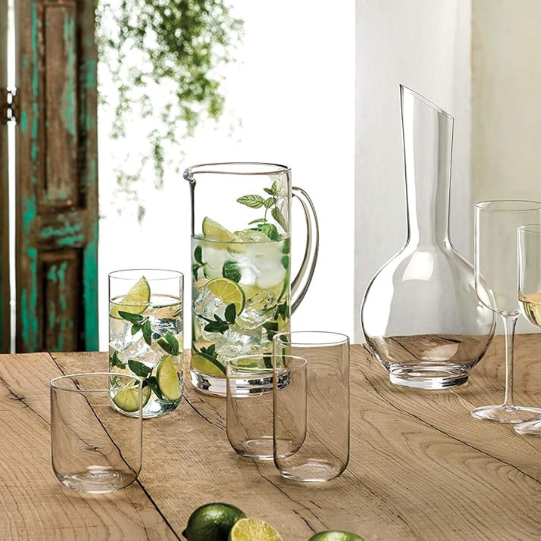 On a wooden table, a Luigi Bormioli glass pitcher with water, ice, lime slices, and mint sits beside empty wine glasses, limes, a decanter, and an oversized Sublime Beverage Glass from the Luigi Bormioli Sublime Beverage Glass Set of 4. Sunlit greenery glows behind.