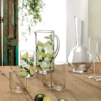 On a wooden table, a Luigi Bormioli glass pitcher with water, ice, lime slices, and mint sits beside empty wine glasses, limes, a decanter, and an oversized Sublime Beverage Glass from the Luigi Bormioli Sublime Beverage Glass Set of 4. Sunlit greenery glows behind.