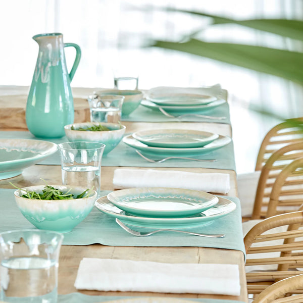 A table set with Taormina Aqua Dinnerware, featuring aqua-colored glazed stoneware plates, bowls, and a matching pitcher, accompanied by glasses and napkins.