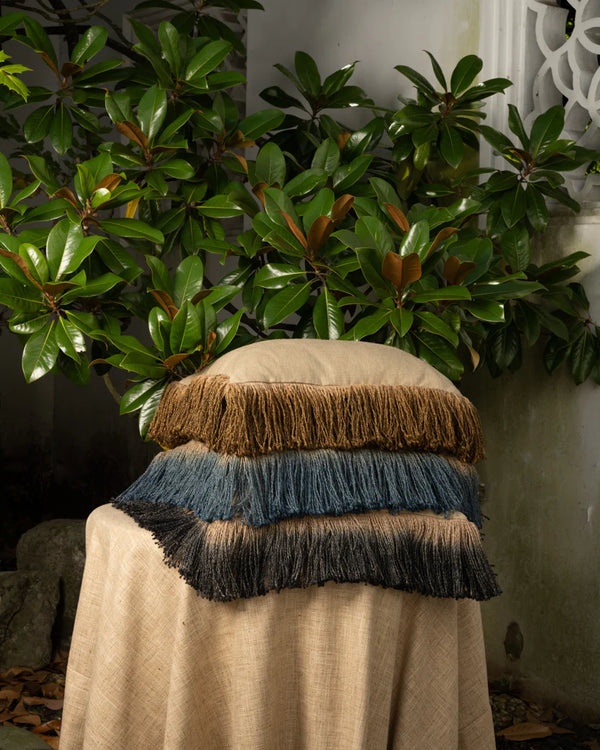 Three Threads Soft Goods Jute Hand Dyed Fringe Pillows in Bronze, each featuring thick fringe borders, are stacked on a cloth-covered chair outdoors, surrounded by green leaves and a white decorative wall.