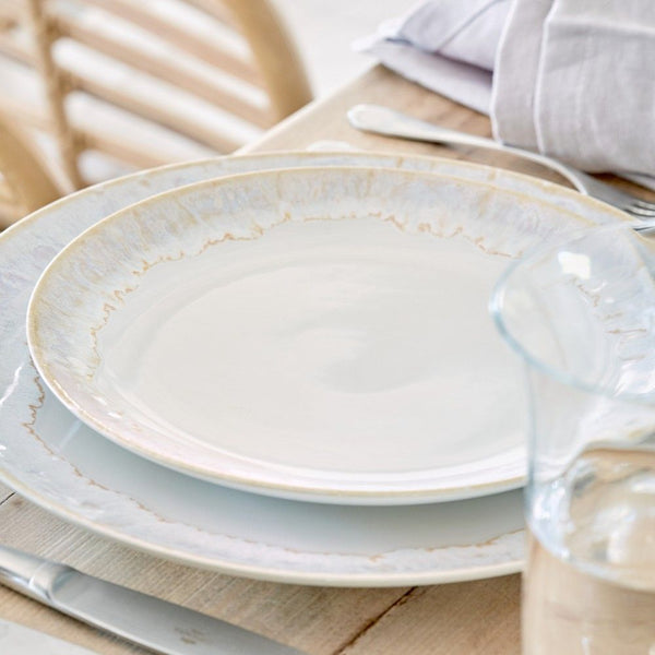 A close-up of a neatly set dining table featuring Casafina's Taormina Dinnerware White, fine stoneware made in Portugal, with a large ceramic plate, knife, fork, glass of water, and folded linen napkin on a light wooden surface.