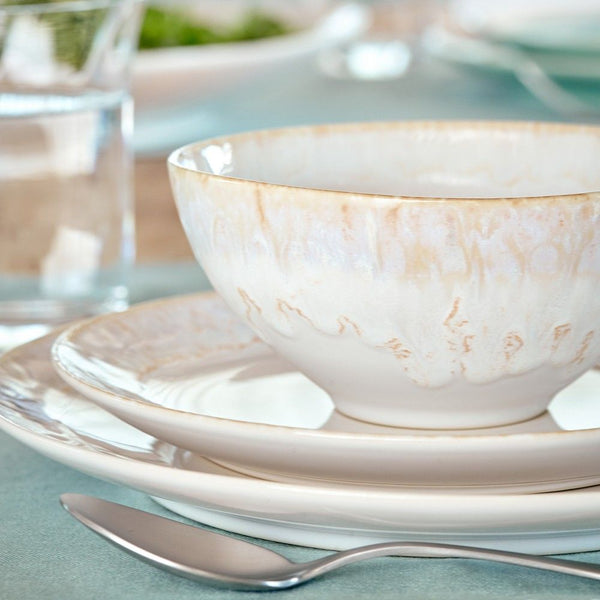 A close-up of Casafina's Taormina Dinnerware White ceramic bowl with a cream and beige pattern, stacked on matching plates. Made in Portugal, it sits beside a spoon and glass of water on a light blue tablecloth.