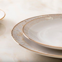 Close-up of two stacked Casafina/Costa Nova Taormina Dinnerware White & Gold ceramic plates with light gray glaze and hand-painted 18-karat gold rim, on a cream marble surface. Part of a third plate is visible. Made in Portugal.