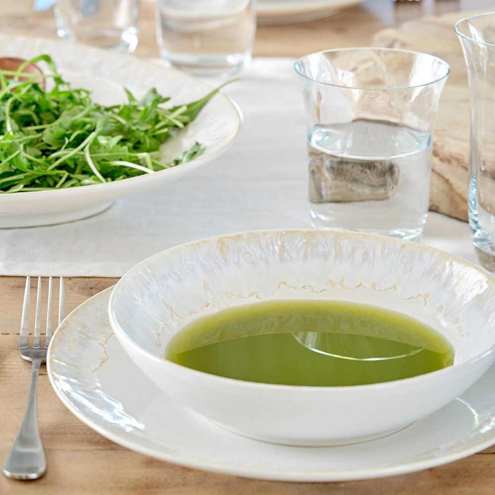 A bowl of green soup sits on the Casafina Taormina Dinnerware White plate with a fork beside it. In the background, a leafy green salad and glass of water rest on the wooden table, showcasing fine stoneware made in Portugal.