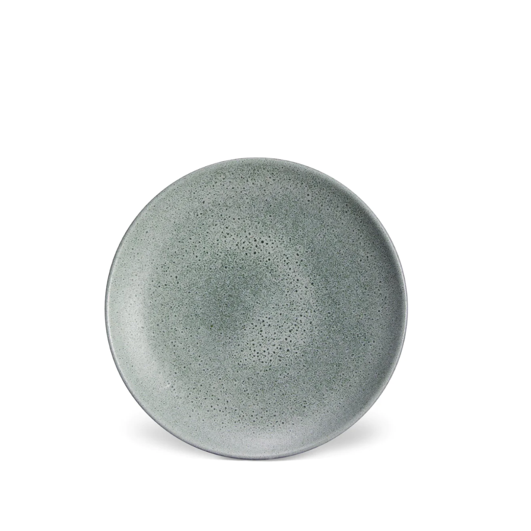 A round, gray ceramic plate with a speckled texture from above on a white background showcases the handcrafted Terra Seafoam Dinnerware by L'Objet, inspired by their signature artisanal style.