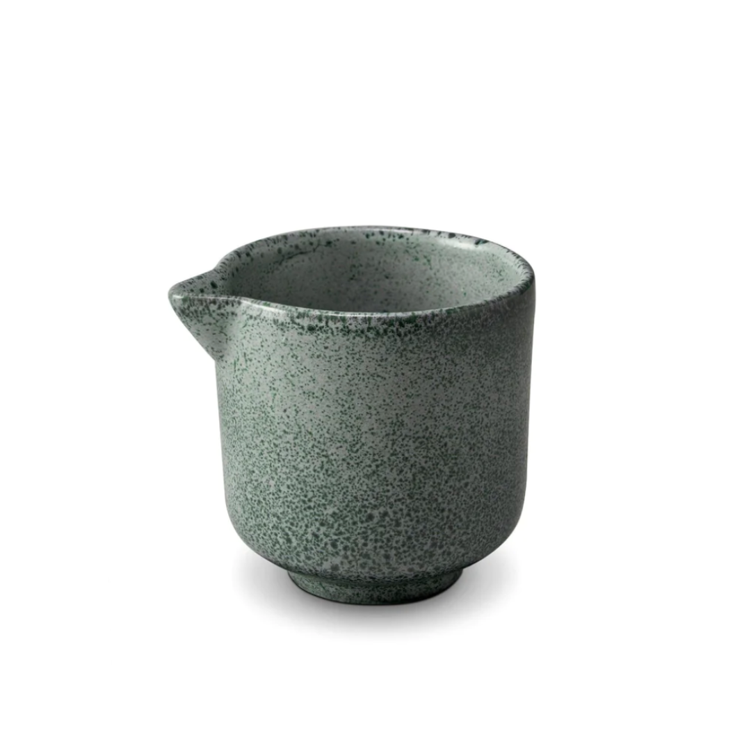 The Terra Seafoam Dinnerware by L'Objet is a small, green speckled ceramic cup with a pouring spout and flared rim, reminiscent of handcrafted pieces, displayed on a white background.