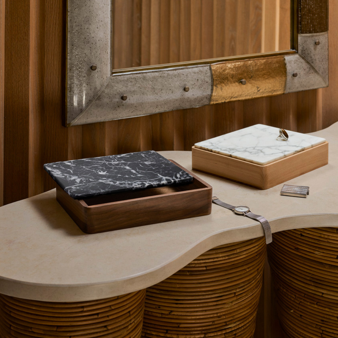 Two Greg Natale Terreno Boxes, one with a black marble lid and one with a white marble lid featuring a ring, sit on a curved light counter beneath a rustic mirror. Nearby rest a watch and small striped item.
