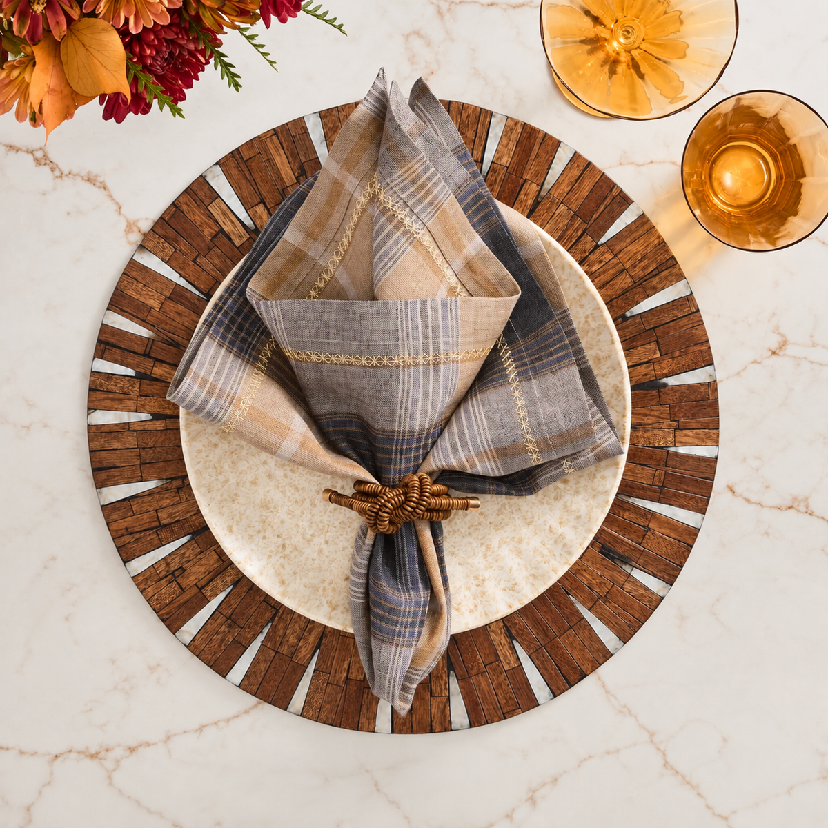 A table setting featuring a round plate, a plaid napkin held by the Kim Seybert Coil Napkin Ring Brown (Set of 4), a wood-patterned charger, two amber glasses, and flowers arranged on a marble surface.