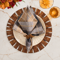 A table setting featuring a round plate, a plaid napkin held by the Kim Seybert Coil Napkin Ring Brown (Set of 4), a wood-patterned charger, two amber glasses, and flowers arranged on a marble surface.