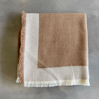Alicia Peru Home’s Eyelash Alpaca Throw, featuring brown and cream tones with a geometric border and fringe edges, is shown folded on a gray surface.