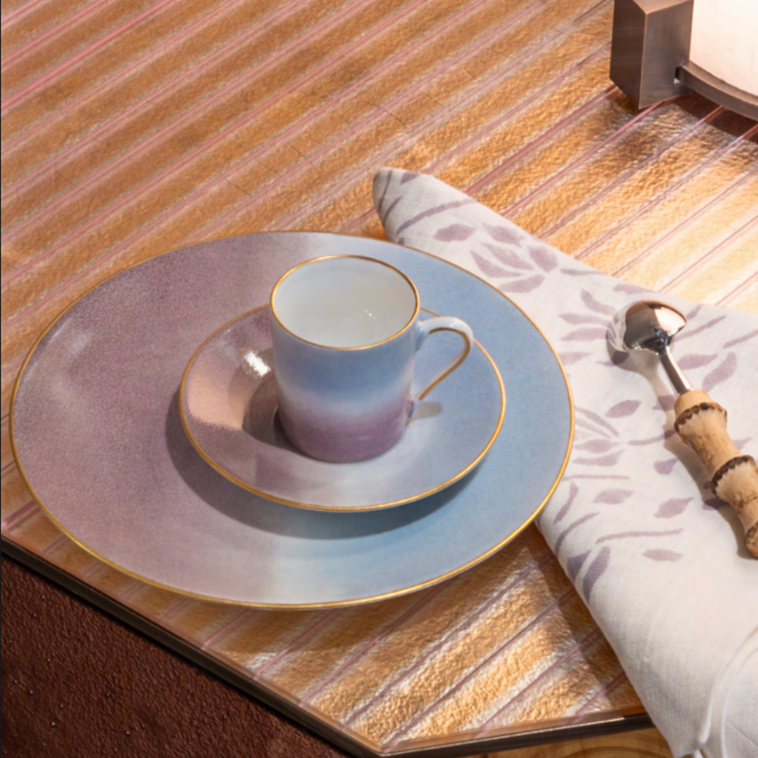 A Marie Daâge Tie Dye Platinum cup and saucer, both hand-painted, rest on a matching dinner plate with platinum rims by Marie Daâge, beside a floral napkin and spoon atop a striped tabletop.