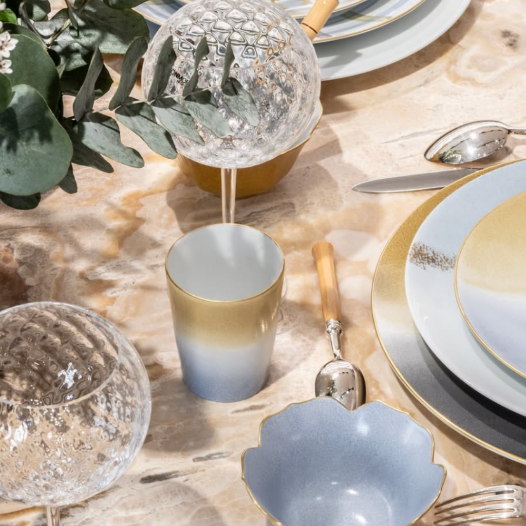 A marble table set with Marie Daage Tie Dye Platinum Dinnerware by Marie Daâge, featuring pastel blue, gold, and white plates, a matching cup, gold utensils, textured glass spheres, green leaves, and a scalloped-edge bowl in soft sunlight.