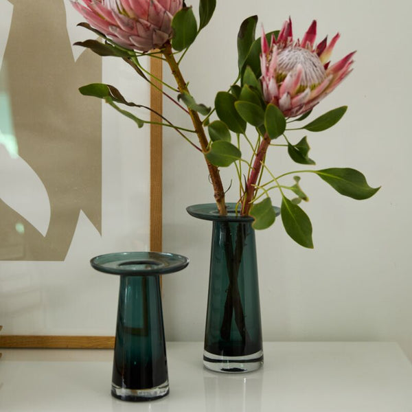 Two Accent Decor Topple Vase Green pieces, one short and one tall, are displayed on a white surface as coffee table decor. The tall vase holds two pink protea flowers with green leaves. An abstract framed art piece hangs in the background.