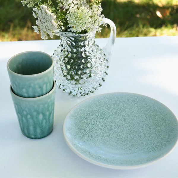 Two stacked sage green Tourron Dinnerware Eucalyptus tumblers rest beside a matching speckled plate and a glass pitcher filled with white flowers.