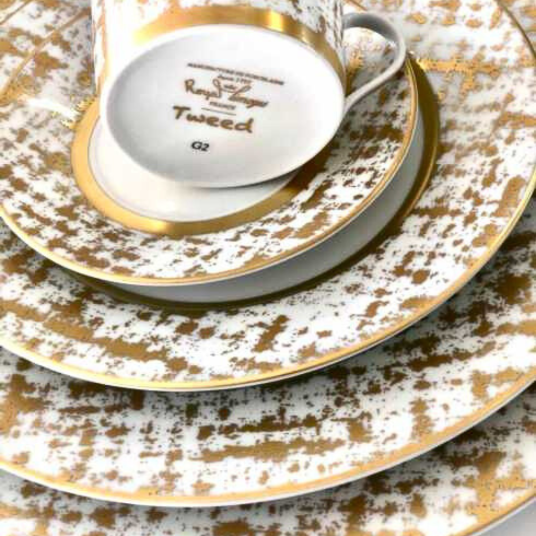 A close-up of stacked Tweed White & Gold Dinnerware by Royal Limoges—white porcelain plates and an upside-down teacup with gold patterns and rims, evoking the elegance of classic French style.