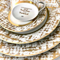 A close-up of stacked Tweed White & Gold Dinnerware by Royal Limoges—white porcelain plates and an upside-down teacup with gold patterns and rims, evoking the elegance of classic French style.