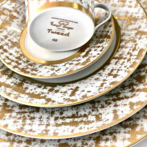 A close-up of stacked Tweed White & Gold Dinnerware by Royal Limoges—white porcelain plates and an upside-down teacup with gold patterns and rims, evoking the elegance of classic French style.