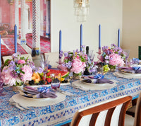 A dining table set with blue and purple candles, floral centerpieces, fruit, and elegant place settings on a blue-patterned tablecloth. Four chairs are visible along one side of the table, while each setting features a Tailored Placemat s/4 - White by Kim Seybert that adds a touch of contemporary style to the inviting arrangement.