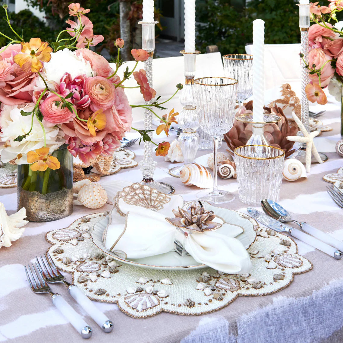 Elegant outdoor table features coastal details: seashell and starfish accents, crystal glassware, gold-rimmed plates, silver cutlery, white candles, florals in pink, peach and white, with Kim Seybert Beachcomber Napkin Ring Set of 4 completing the look.