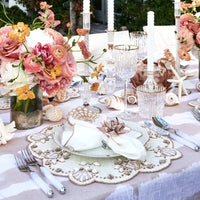 Elegant outdoor table features coastal details: seashell and starfish accents, crystal glassware, gold-rimmed plates, silver cutlery, white candles, florals in pink, peach and white, with Kim Seybert Beachcomber Napkin Ring Set of 4 completing the look.