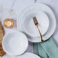 A place setting featuring Vietri's Aria Dinnerware, including a handcrafted stoneware dinner plate, white ceramic bowls, a gold fork in a bowl, green cloth napkin, lit candle in glass holder, and wooden board on marble.