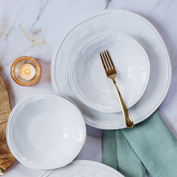 A place setting featuring Vietri's Aria Dinnerware, including a handcrafted stoneware dinner plate, white ceramic bowls, a gold fork in a bowl, green cloth napkin, lit candle in glass holder, and wooden board on marble.