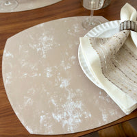 A close-up of a luxe dining table set with Bodrum's NEW Velvet Elliptic Square Placemat, a white scalloped plate, a folded cream napkin with patterned stripe in a silver ring, and a glass holding a beige napkin.