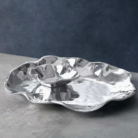The Beatriz Ball Vento Bergamo Oven Platter & Bowl, crafted from shiny aluminum alloy with a wavy edge, features a matching smaller bowl resting atop the platter and is displayed on a gray surface against a dark blue background.