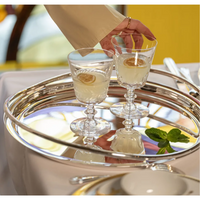 A hand reaches for the Vertigo Silver Plated Round Tray by Christofle, holding two glasses of pale yellow liquid with lemon slices, reflecting the light.