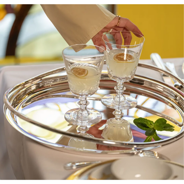 A hand reaches for the Vertigo Silver Plated Round Tray by Christofle, holding two glasses of pale yellow liquid with lemon slices, reflecting the light.