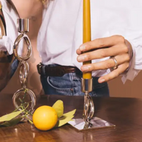 A person in a white shirt and jeans places a yellow candle into the Christofle Vertigo Silver Plated Candlestick on a wooden table beside another modern candlestick, a lemon, and green leaves.
